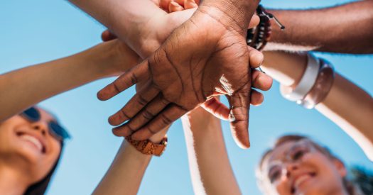bottom view of multiracial young friends holding hands together with blue sky on backdrop