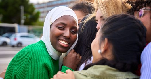 Portrait of black Muslim woman looking smiling at camera surrounded in an embrace by multiracial female people. African girl student in white hijab happy with her friends standing together outdoor